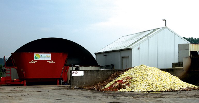 View of the Joluwa biogas plant. To the left the red receiving bin with the hydrolysis tank, the HYFAD unit is inside the shed. In the foreground the feedstock chicory roots and endive leaves alongside the solid digestate discharge. Further to the right, out of view, is the CHP unit and the post digester.